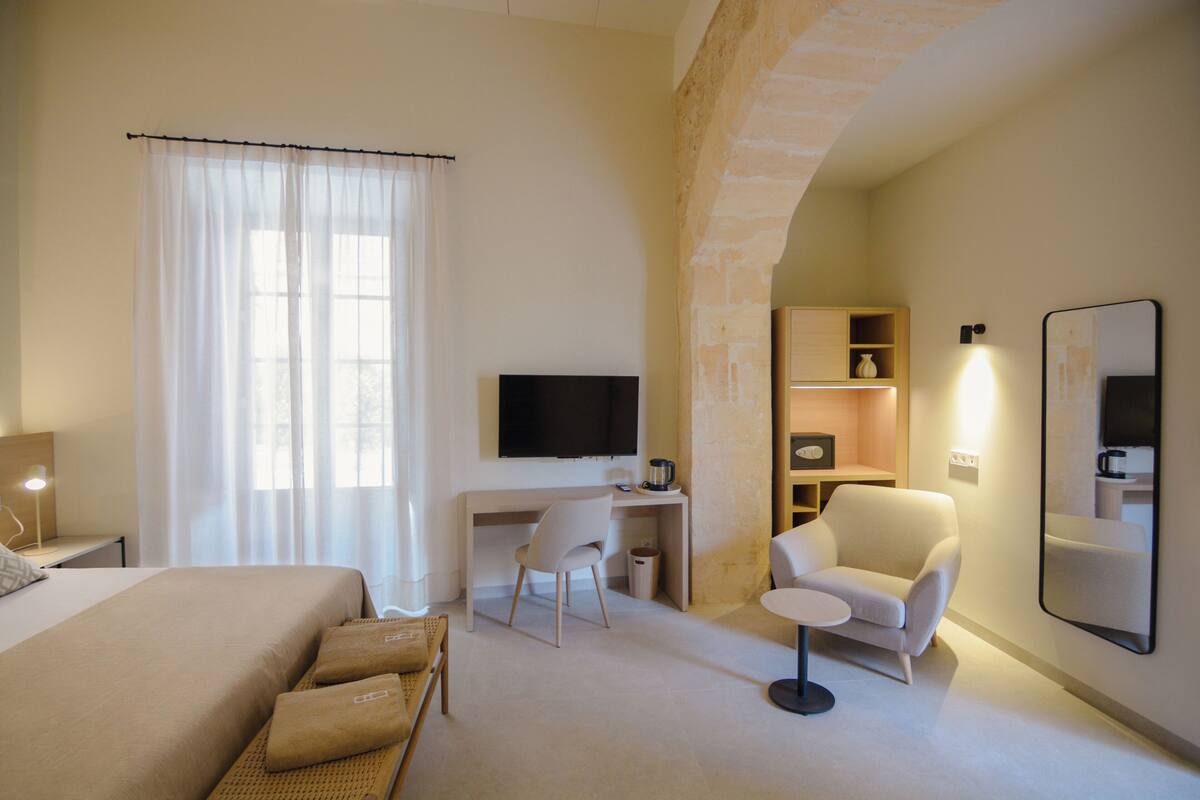 A hotel room with a bed, a desk, a chair, a TV, a mirror, and a small table.