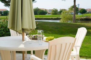 A round outdoor table with a green umbrella, two chairs, a pitcher, glasses, and a plate of grapes. - Bon marché ! Maison avec terrasse privée | + Golf (Saint Gilles Croix de Vie)
