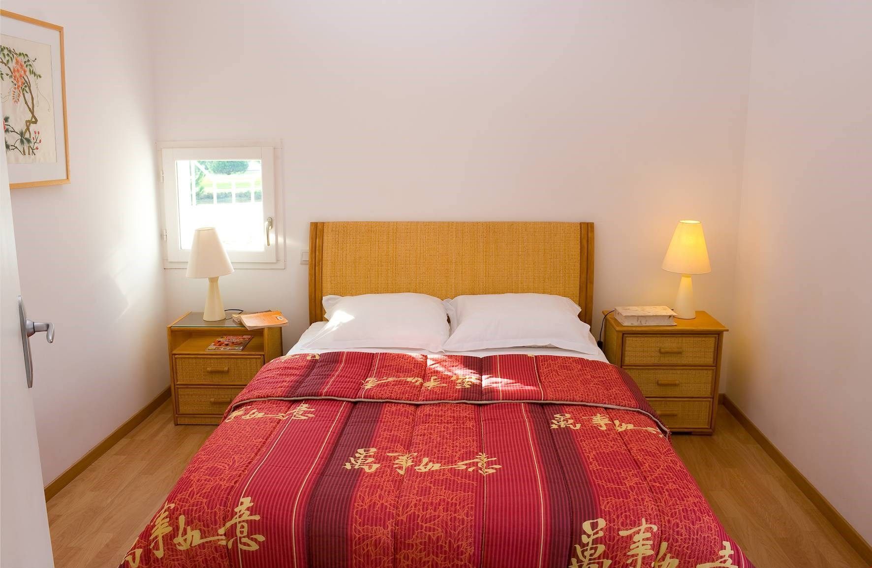 A neatly made bed with a red and gold patterned bedspread, two bedside tables with lamps, a window with blinds, and a framed picture on the wall.