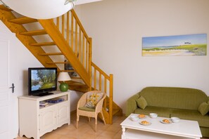 Ein Wohnzimmer mit einer Holztreppe, einem Fernseher, einem grünen Sofa, einem Korbstuhl und einem Gemälde mit Strandmotiv.