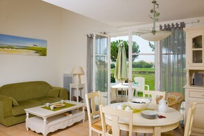 Ein gemütliches Wohnzimmer mit einem grünen Sofa, einem Esstisch mit Stühlen und Blick auf einen Garten durch große Fenster.