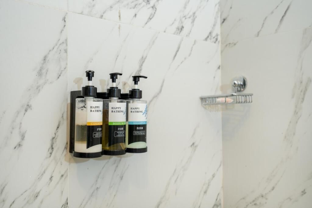 Un baño con paredes de mármol, ducha y tres botellas de jabón corporal Happy Bathing.
