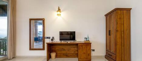 Una habitación con un escritorio de madera, televisor, espejo, armario y ventana con cortinas.