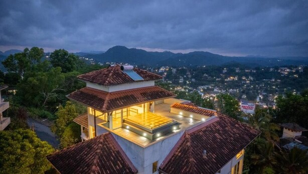 A modern house with a rooftop terrace, surrounded by lush greenery and mountains.