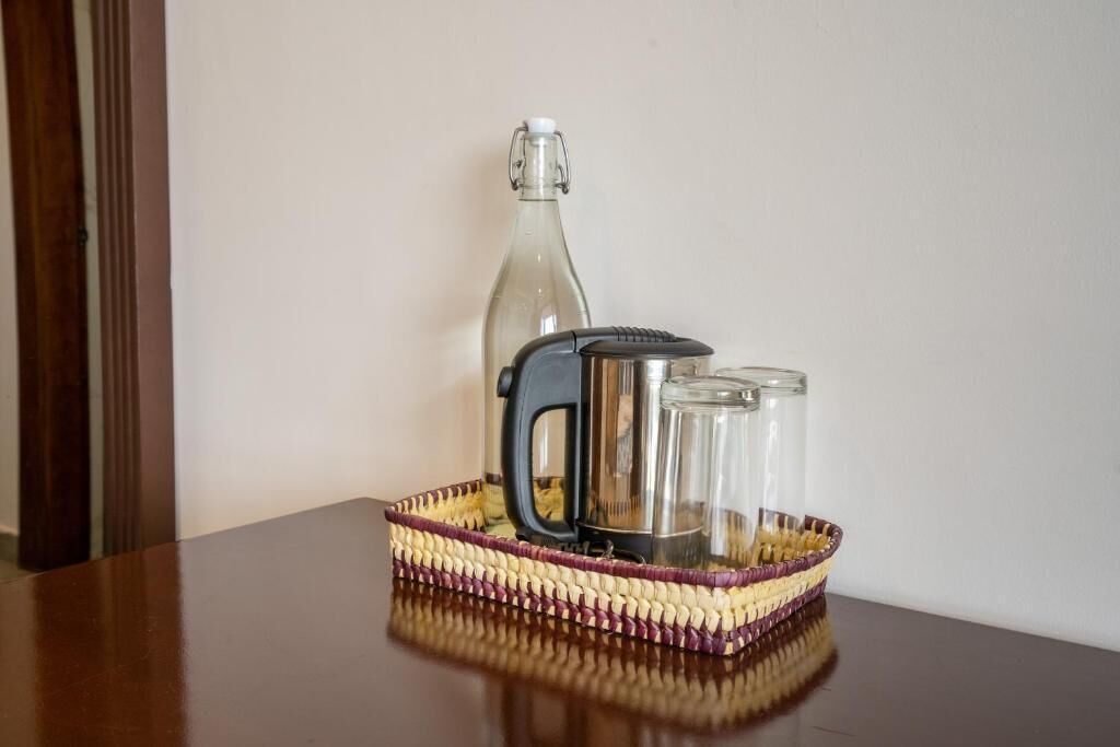 A glass bottle, a kettle, and two glasses on a woven tray on a table.