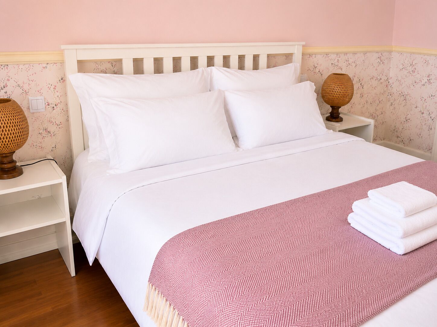 Un letto rifatto con lenzuola bianche, una coperta rosa fantasia e una pila di asciugamani bianchi su un lato.