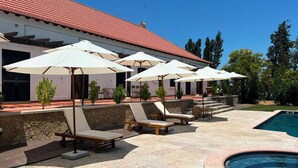 Un espace aménagé au bord de la piscine, avec des chaises longues et des parasols, un mur en pierre et un bâtiment surmonté d’un toit en tuiles rouges.
