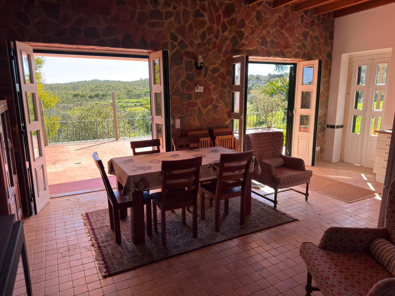 A dining area with a table and chairs, a patterned rug, and a view of a green landscape through open doors.