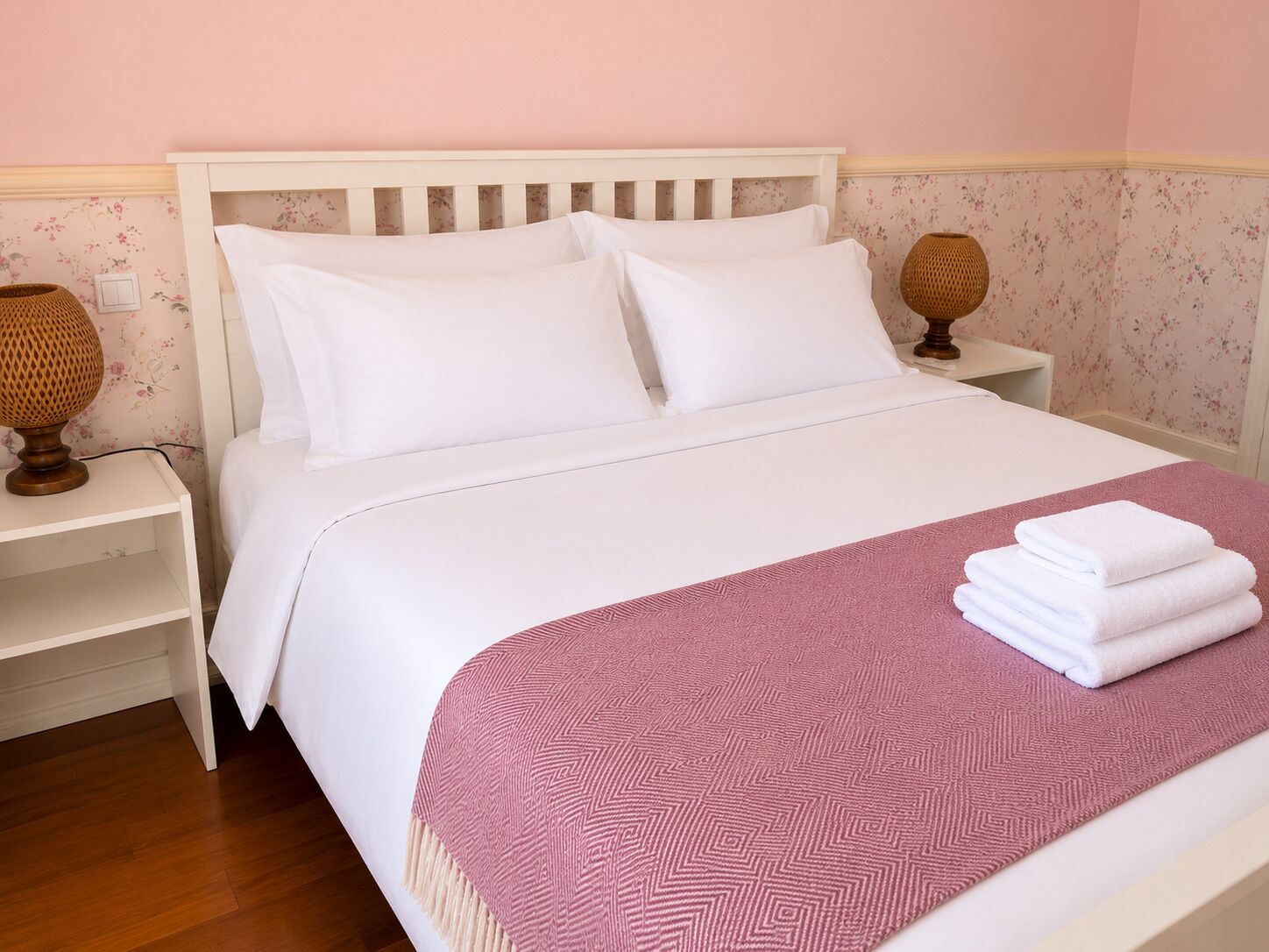 Una cama impecablemente tendida con sábanas blancas y un plaid rosa estampado, dos lámparas de noche y una pared cubierta de papel tapiz floral.