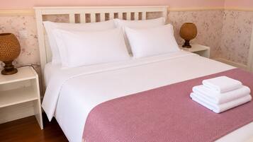 Una cama impecablemente tendida con sábanas blancas y un plaid rosa estampado, dos lámparas de noche y una pared cubierta de papel tapiz floral.
