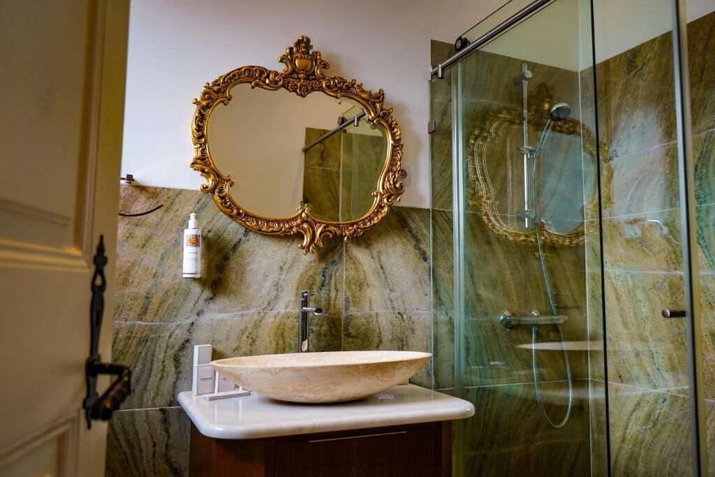 Un baño con pared de mármol, un espejo con marco dorado, un lavamanos tipo cuenco y una ducha con mampara de vidrio.