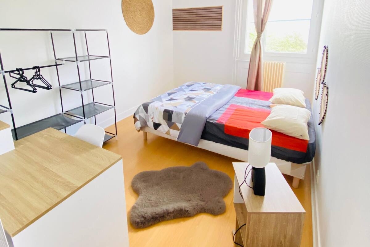 A modern bedroom with a bed, a desk, a hanging light fixture, and a window with curtains.