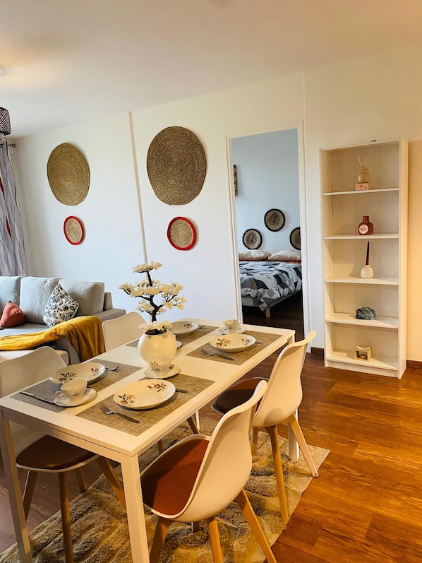 A dining area with a table set for four, chairs, a vase with flowers, and a bookshelf in the background.