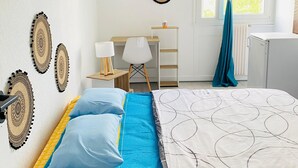 Ein Schlafzimmer mit Bett, Schreibtisch, Stuhl, Lampe und einem fenster mit Vorhängen.
