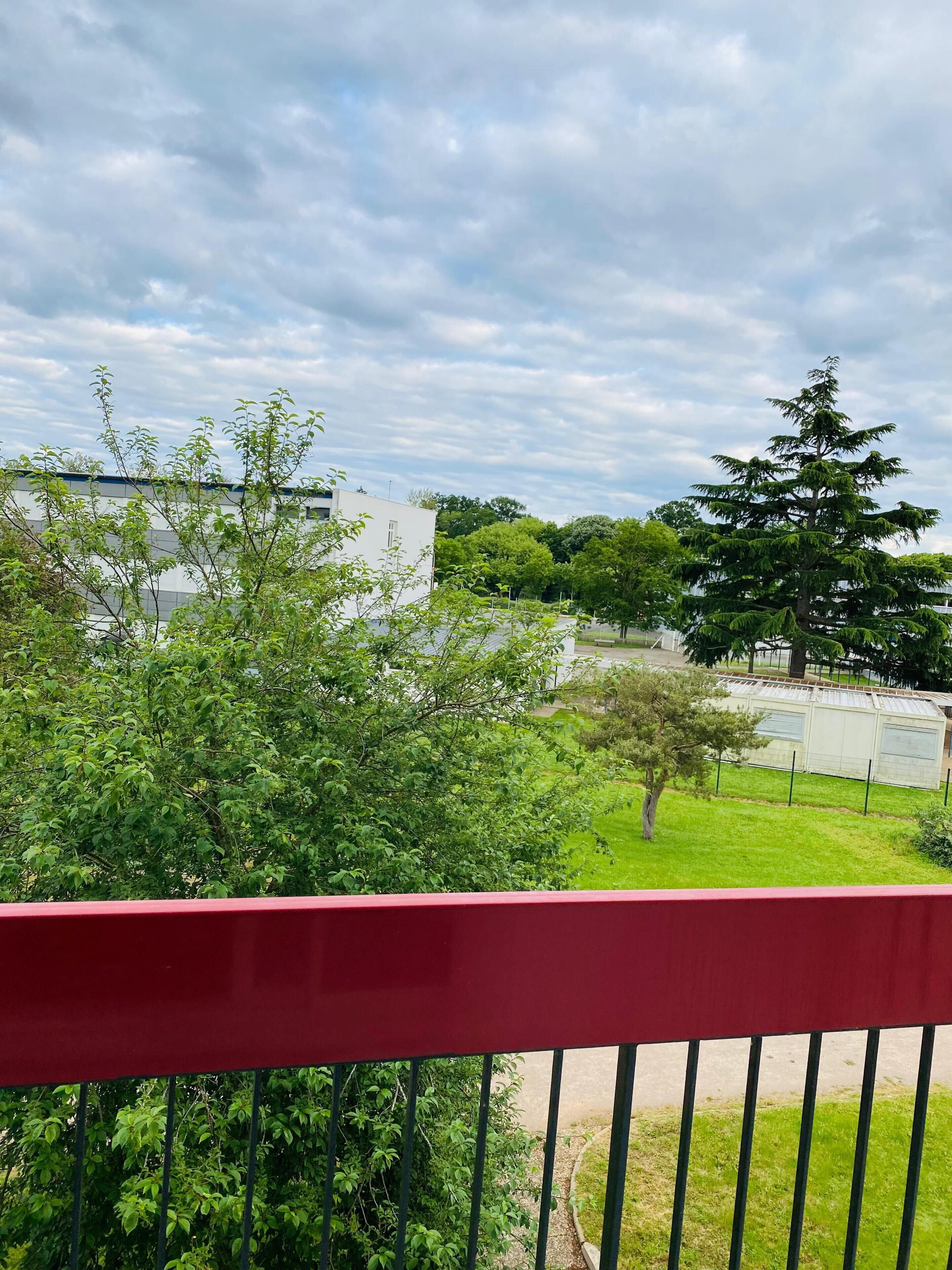 Un balcon avec une balustrade rouge donnant sur une pelouse, un grand arbre et un bâtiment à la façade blanche.