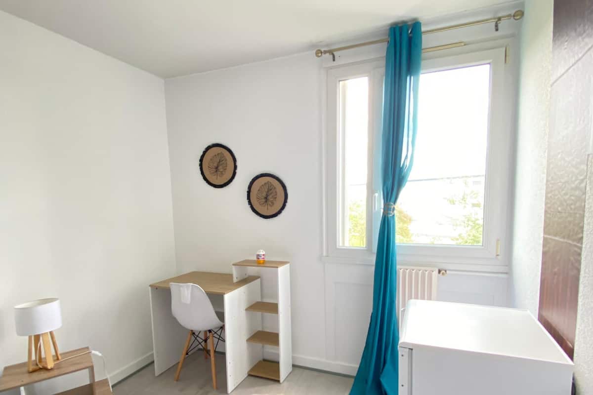 A compact bedroom with a bed, a desk, a chair, a radiator, and a window with curtains.