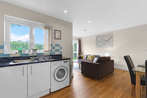 Eine moderne Küche mit Waschmaschine, eine Essecke mit Glastisch und ein Wohnzimmer mit Sofa.