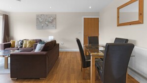 A modern living room with a sofa, a dining table, and chairs. There is a large mirror on the wall and a map artwork. - Heathrow Living 2BDR 2BA Apt by 360 Stays - Apt 02 (Stanwell)