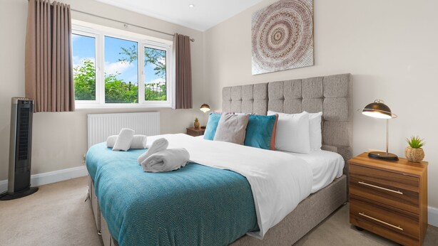 A bedroom with a bed, a bedside table with a lamp, a radiator, and a window with curtains. - Heathrow Living 2BDR 2BA Apt by 360 Stays - Apt 02 (Stanwell)