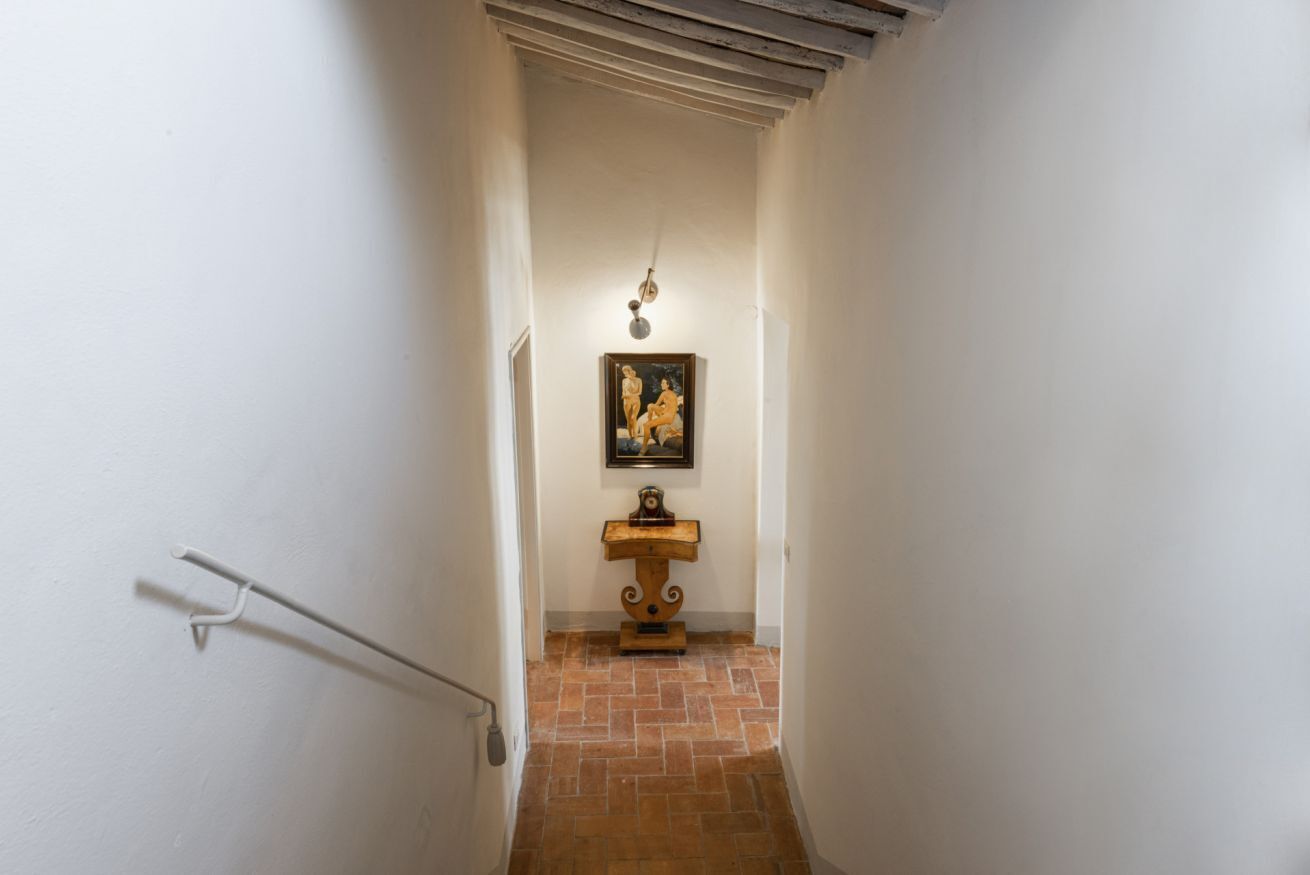 A narrow hallway with white walls, a tiled floor, a wooden stand with a painting, and a light fixture.