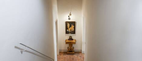 A narrow hallway with white walls, a tiled floor, a wooden stand with a painting, and a light fixture.