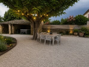 Une terrasse en gravier avec une table et des chaises, un espace barbecue en pierre et un arbre décoré de guirlandes lumineuses.