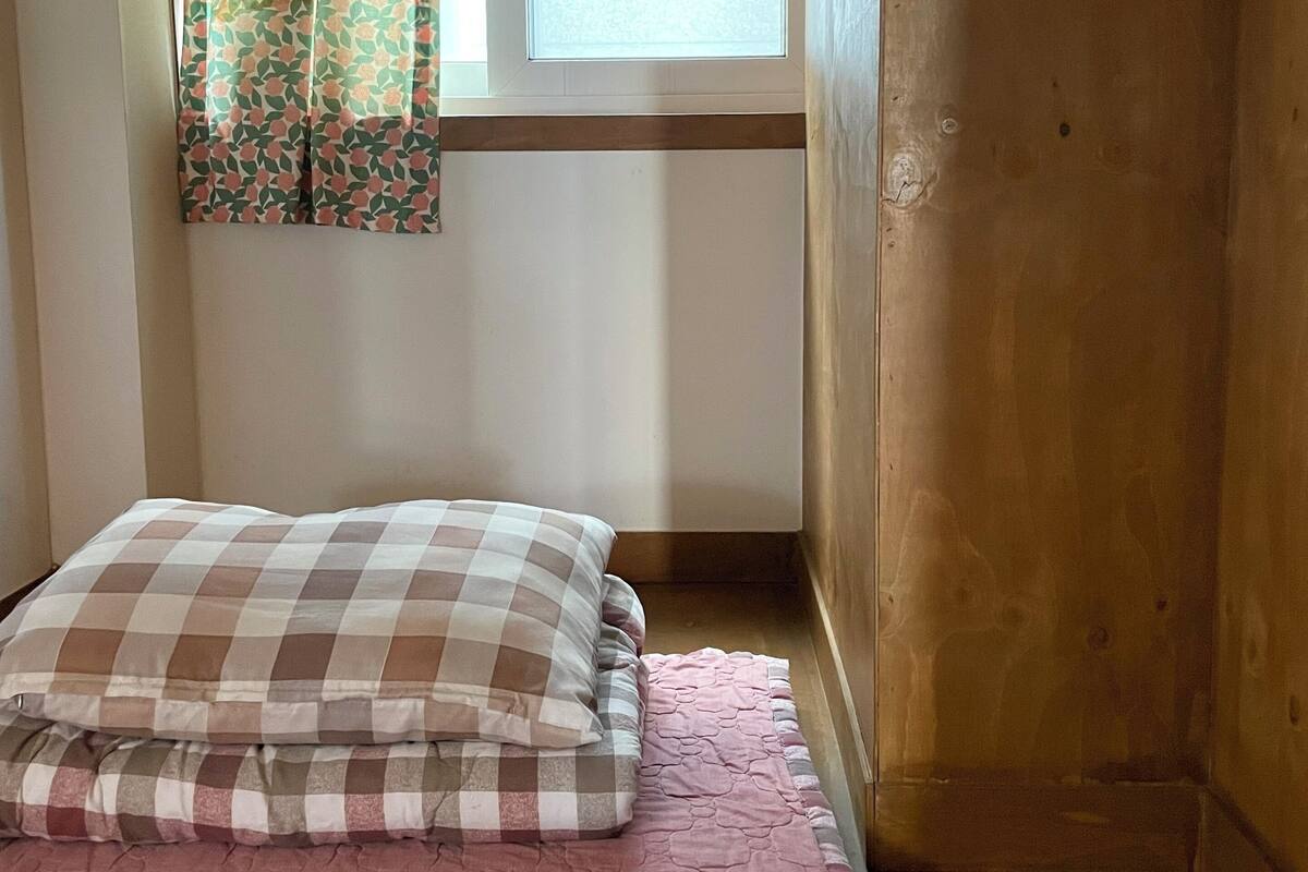 A small, enclosed room with a bed, a window with patterned curtains, and a wooden ceiling.