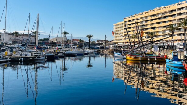 A marina with boats docked, a hotel building in the background, and clear skies.