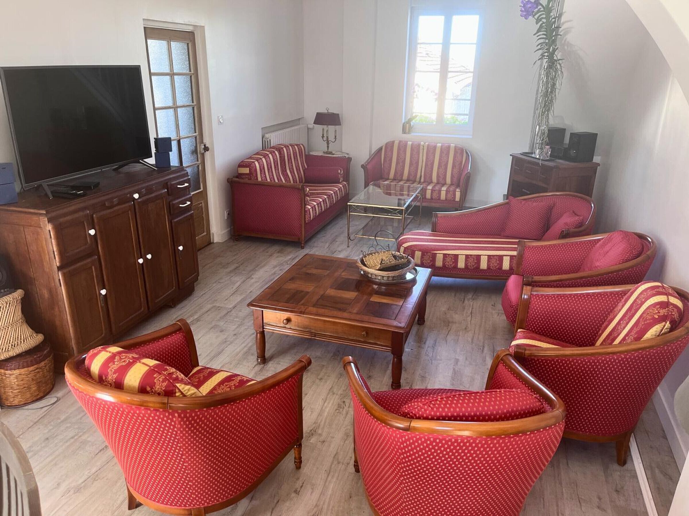 Una sala de estar con televisor, aparador de madera, sillones tapizados en rojo, una mesa de centro y un ventanal con luz natural.