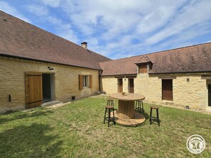 A courtyard with a round wooden table and stools, stone buildings with tiled roofs, and a clear sky. - House in Périgord, parking, pets allowed - 4 guests (Campagne)