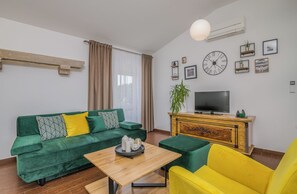 Uma sala de estar com um sofá verde, uma poltrona amarela, uma mesa de centro de madeira, uma TV em um armário de madeira e um relógio de parede grande.
