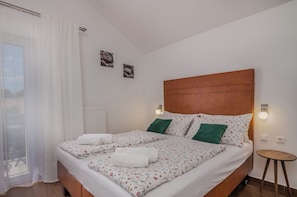 Uma cama de casal com roupa de cama floral, dois travesseiros verdes, uma cabeceira de madeira, uma pequena mesa redonda e duas luminárias fixadas na parede.