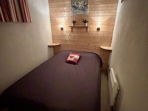 A small, cozy room with a wooden wall, a bed with a purple bedspread, two bedside tables, and a small shelf with plants. - Apartment with parking - 4 guests (Saint-Lary-Soulan)