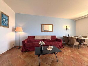 A living room with a red sofa, a black coffee table with a vase and magazine, a lamp, a dining table with chairs, and a wall-mounted mirror. - Apartment stunning sea view - 4 guests (Saint-Cyr-sur-Mer)