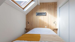 Een slaapkamer met een schuine kap, houten paneelwanden, een bed met wit beddengoed, een gele plaid, een wandklok en een dakraam.