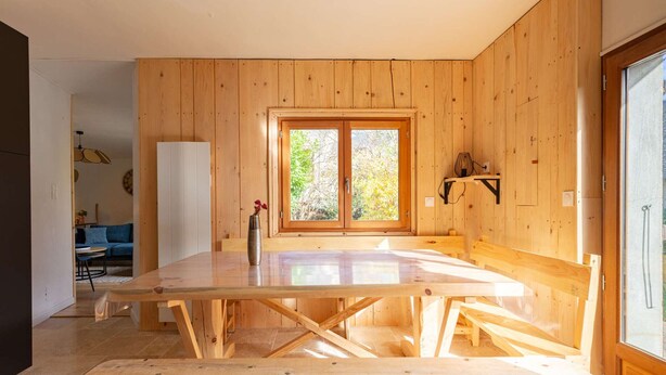 Een houten interieur met een grote tafel en banken, een deur met ruiten en uitzicht op groen.