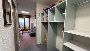 Ein schmaler Flur mit einem Bett, einem Sessel und einem Kleiderschrank mit offenen Regalen.