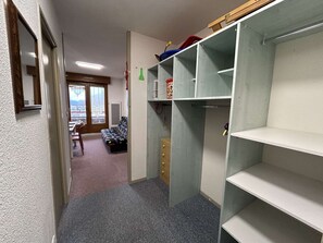 Ein schmaler Flur mit einem Bett, einem Sessel und einem Kleiderschrank mit offenen Regalen.