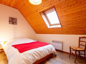 Um quarto com teto de madeira inclinado, um claraboia, uma cama com roupa de cama branca e um cobertor vermelho, uma cadeira de madeira e um radiador.