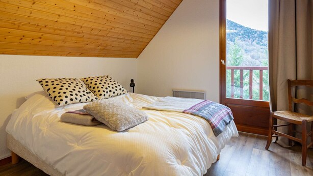 A bedroom with a wooden ceiling, a bed with white bedding, two polka-dotted pillows, a blanket, a chair, and a view of greenery outside.