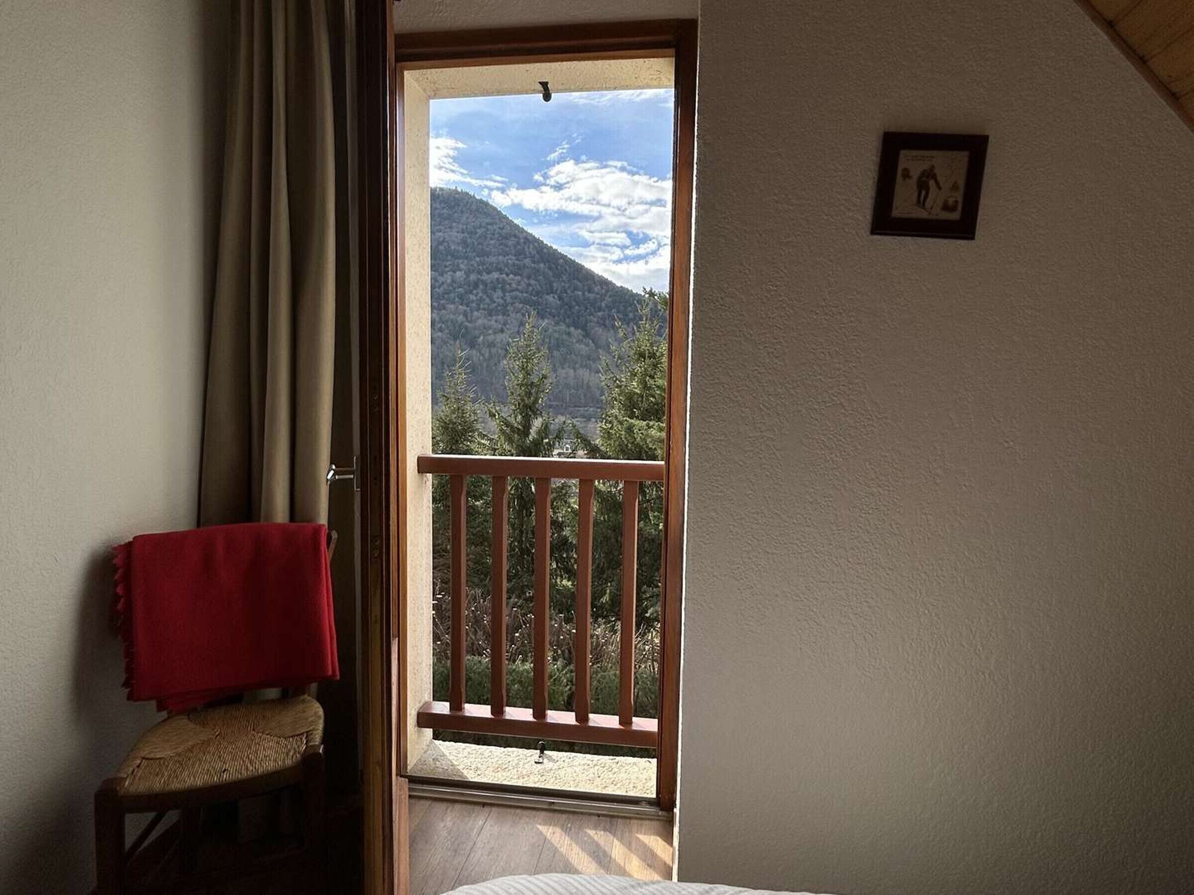 A room with a wooden chair, a red towel, a balcony with a wooden railing, and a mountain view.