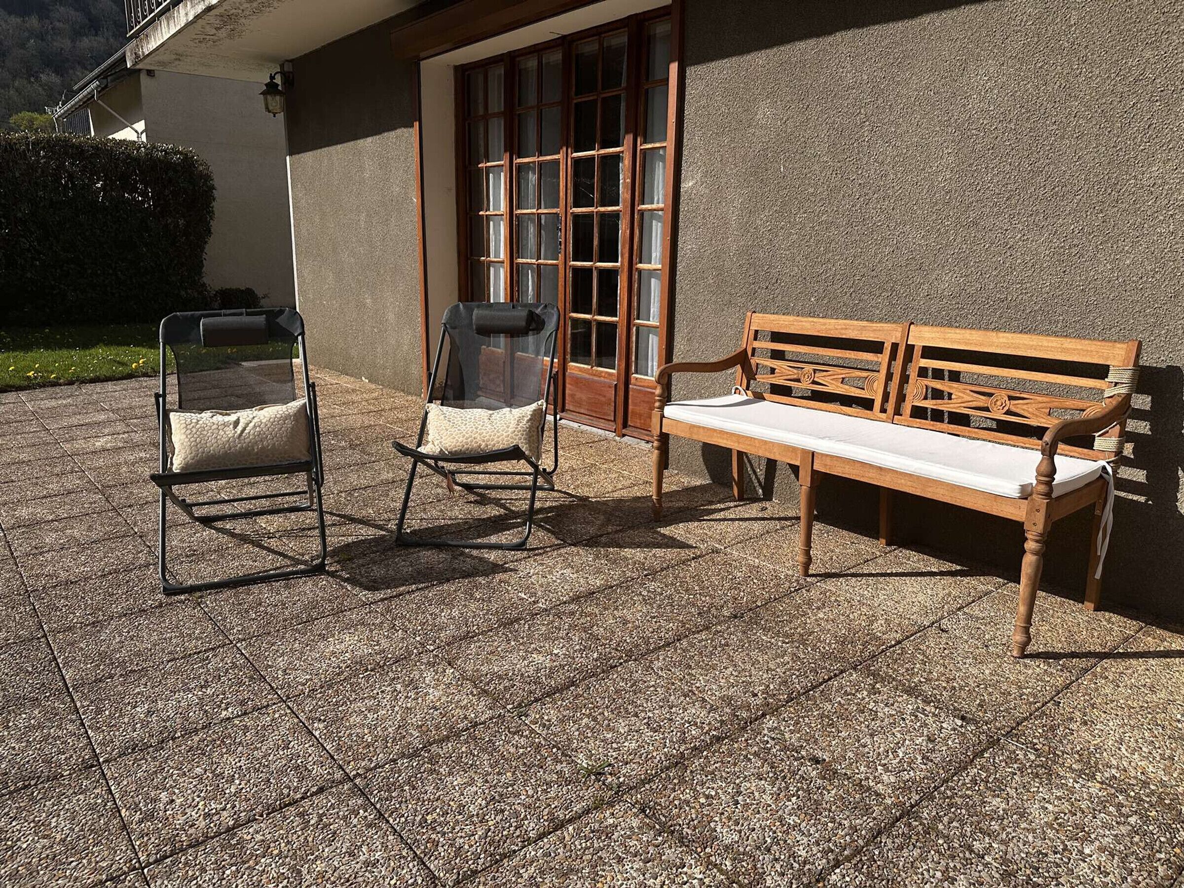 Un espace extérieur aménagé avec un banc et une chaise, l’entrée d’un bâtiment et une haie.