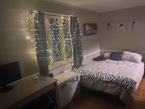 A bedroom with a bed, a desk, a window with string lights, and a painting on the wall.