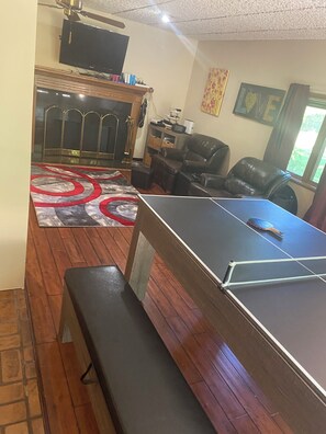 Quarto com mesa de pingue-pongue, um banco, lareira, televisão e janela com cortinas.