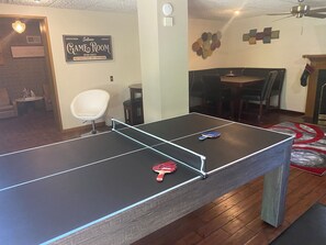 Une table de ping-pong dans une pièce où se trouvent une chaise blanche, une table à manger et un panneau mural indiquant « SALLE DE JEUX ».
