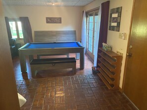 Une table de billard dans une pièce dont le sol est recouvert de briques, avec un banc, une fenêtre avec des rideaux et une porte sur laquelle est fixé un porte-serviettes.