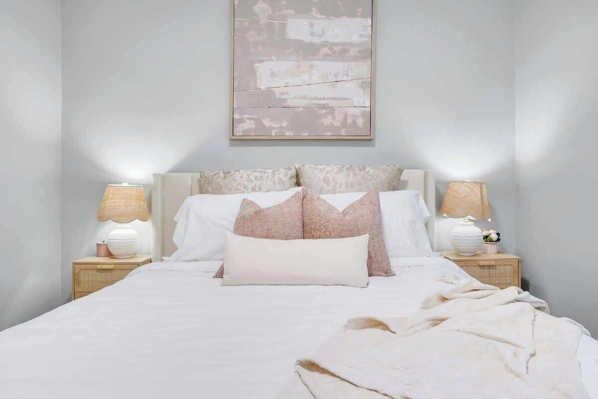 Cozy king bed with layered pillows, soft throw, matching bedside tables, elegant lamps, and delicate floral accents under modern wall art.
