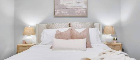 Cozy king bed with layered pillows, soft throw, matching bedside tables, elegant lamps, and delicate floral accents under modern wall art.