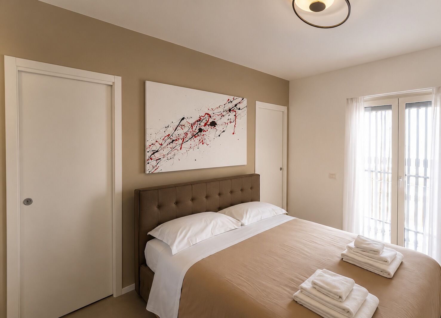 A bedroom with a large bed, a headboard, a painting on the wall, and a window with curtains.