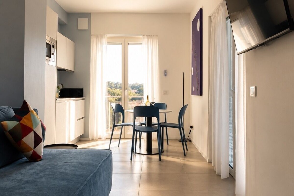 Una sala moderna con un sofá, una mesa de comedor con sillas, un televisor de pantalla plana montado en la pared y una zona de cocina con armarios y un microondas.
